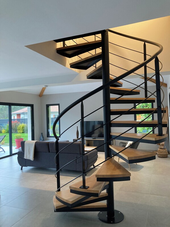 A modern spiral staircase with a minimalist design, a grey sofa, and a TV in a spacious living area with large windows.