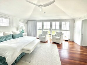 A spacious bedroom with a large bed, two white armchairs, a ceiling fan, and a view through large windows.