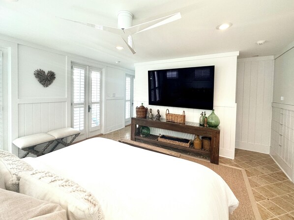A bedroom with a large bed, a TV mounted on the wall, a ceiling fan, and a bench by the window.
