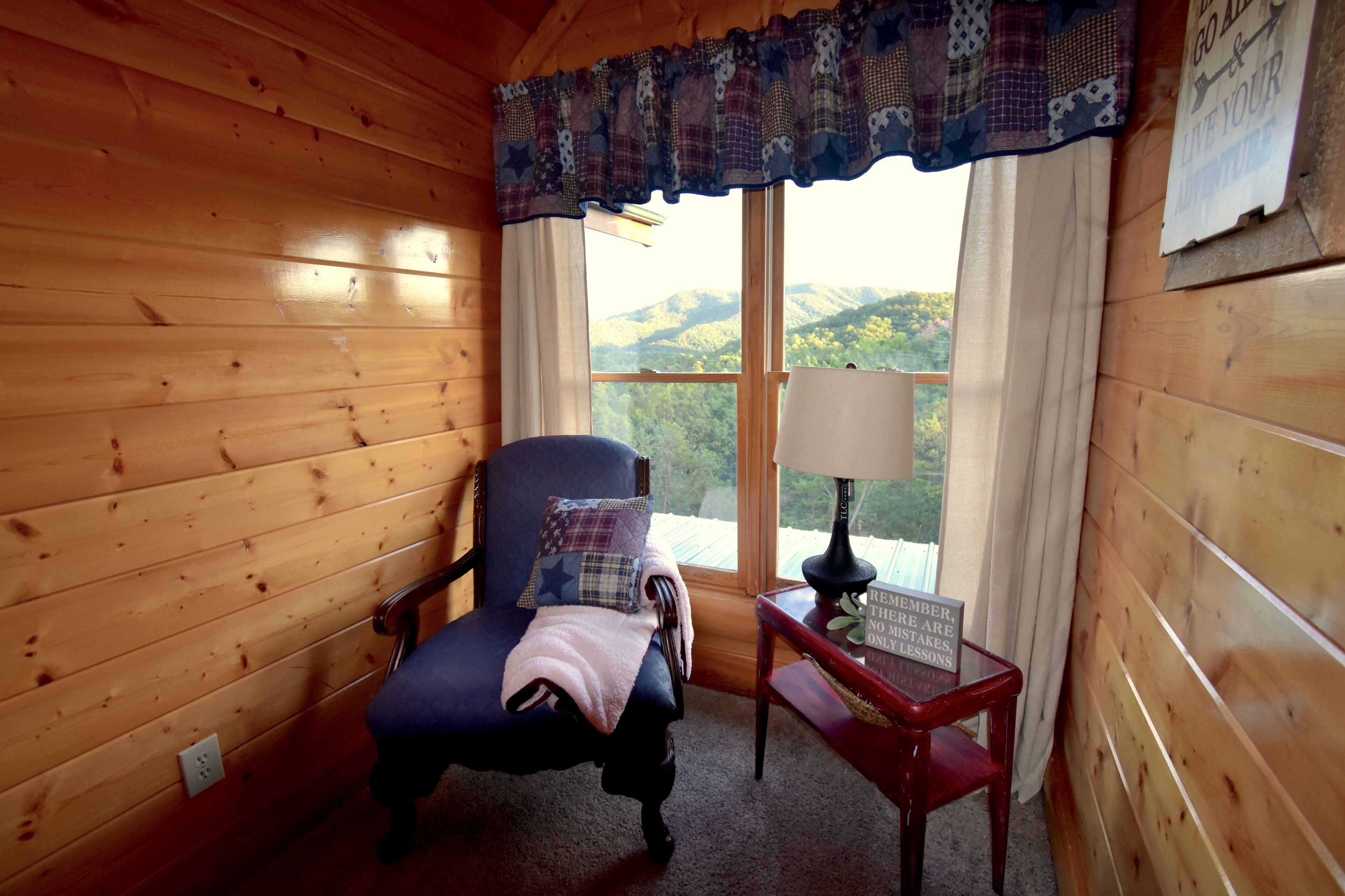 A cozy room with wooden walls, a blue armchair with a star pillow, a small table with a lamp, and a window with a view of green hills.