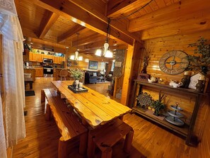 A wooden interior with a long dining table, benches, a kitchen area, and a fireplace with decorative items.