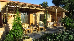Un edificio rústico con una pérgola de madera, mobiliario exterior y un jardín con macetas y flores.