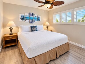 A bedroom with a large bed, two bedside tables with lamps, a ceiling fan, a window with blinds, and a wall art piece.