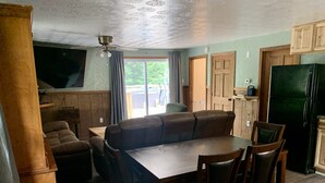A cozy living room with a wooden floor, a large brown leather sofa, a dining table with chairs, a ceiling fan, and a kitchen area with a refrigerator.