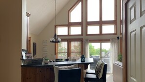 A kitchen with a wooden island, two bar stools, a hanging light fixture, and large windows with blinds.