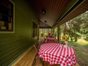 A covered porch with a checkered table and chairs, a window, and a view of trees. - Sunset Lake Cabin in Ripley, NY (Ripley)