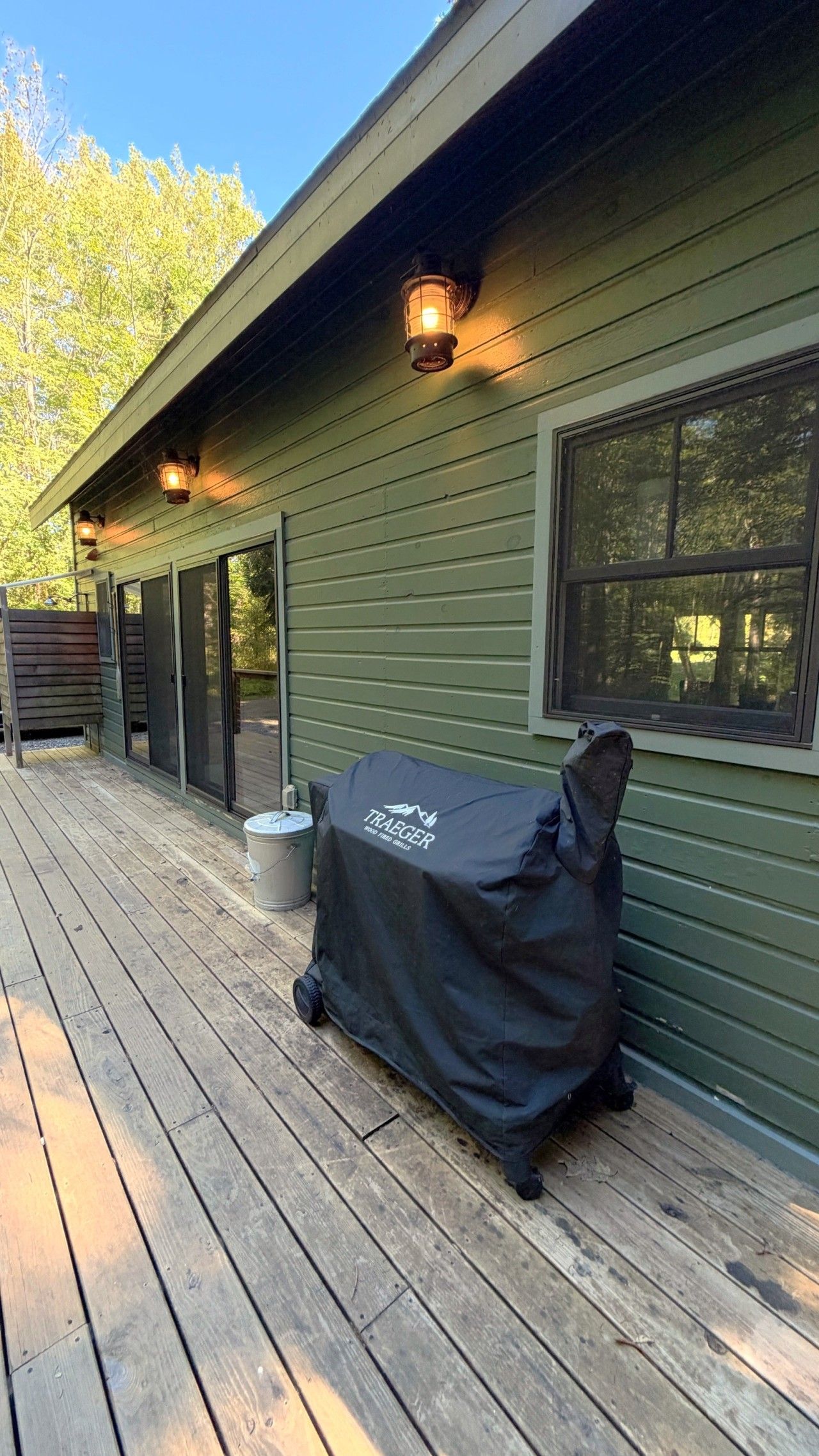 A wooden deck with a grill cover, a trash can, and a house with green siding and a window.