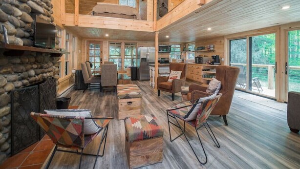 A spacious living area with a stone fireplace, wooden furniture, and large windows offering a view of the outdoors.