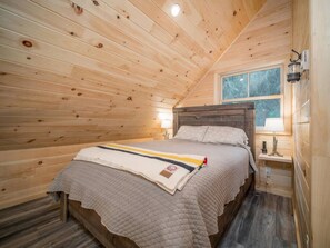 A cozy wooden cabin bedroom with a bed, a nightstand, a lamp, and a window. - Sunset Lake Cabin in Ripley, NY (Ripley)