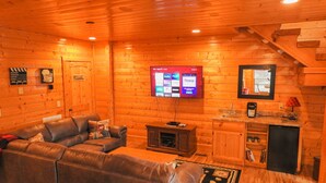 A cozy log cabin interior with a leather sofa, a flat-screen TV mounted on the wall, a small kitchenette, and a wooden staircase. - MTN views! PoolTable, AirHockey, Foosball, HotTub, FirePit; Perfect for FAMILIES (Sevierville)