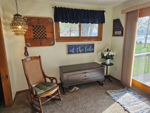 A room with a wooden rocking chair, a chessboard, a sign reading 'Life is better at the lake,' a small table with a potted plant, and a view of a lake through a window.