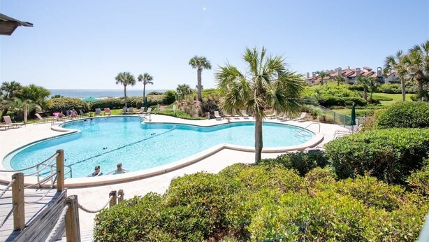 Une piscine à bordures courbes, entourée de palmiers et de chaises longues, offrant une vue sur l’océan et une zone résidentielle en arrière-plan.