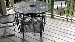 Une table ronde en métal, entourée de chaises, est posée sur une terrasse en bois. Une rambarde en métal entoure l’ensemble, et on aperçoit des arbres et un bâtiment en arrière-plan.