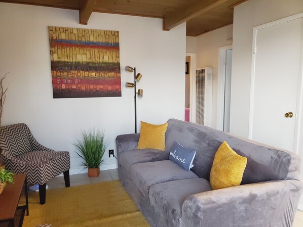 Un salon avec un canapé gris, un fauteuil à motifs, une lampe sur pied et un grand tableau abstrait accroché au mur.
