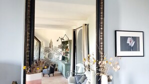 Um quarto com um espelho grande e ricamente decorado sobre um console de mármore preto, uma fotografia emoldurada na parede e uma planta em vaso com flores amarelas.