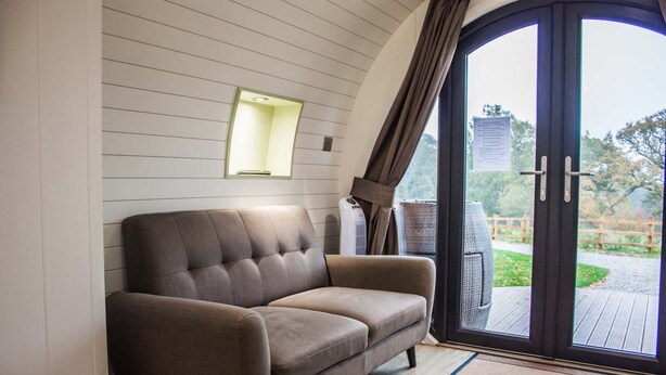 A modern living room with a brown sofa, a wall-mounted light, and a sliding glass door leading to an outdoor area. - Foxtail Glamping Pod - sleeps 2 | Gold Award winning (Pickering)