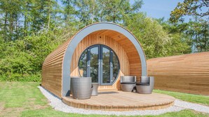 Eine moderne, halbkreisförmige Holzkonstruktion mit Glastür, zwei Korbstühlen und einem kleinen Tisch auf einer Holzterrasse.