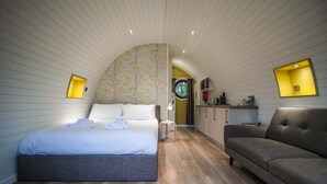 Ein Schlafzimmer mit Bett, einer Couch, einer kleinen Küchenzeile und einem Badezimmer mit rundem Fenster.
