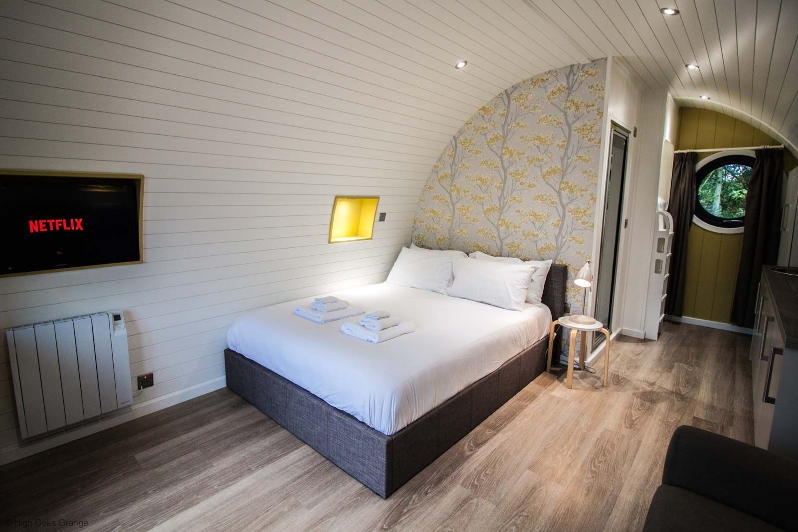 Ein Schlafzimmer mit Bett, einem Fernseher, der 'NETFLIX' zeigt, einer Nachttischlampe und Blick aus dem Fenster.