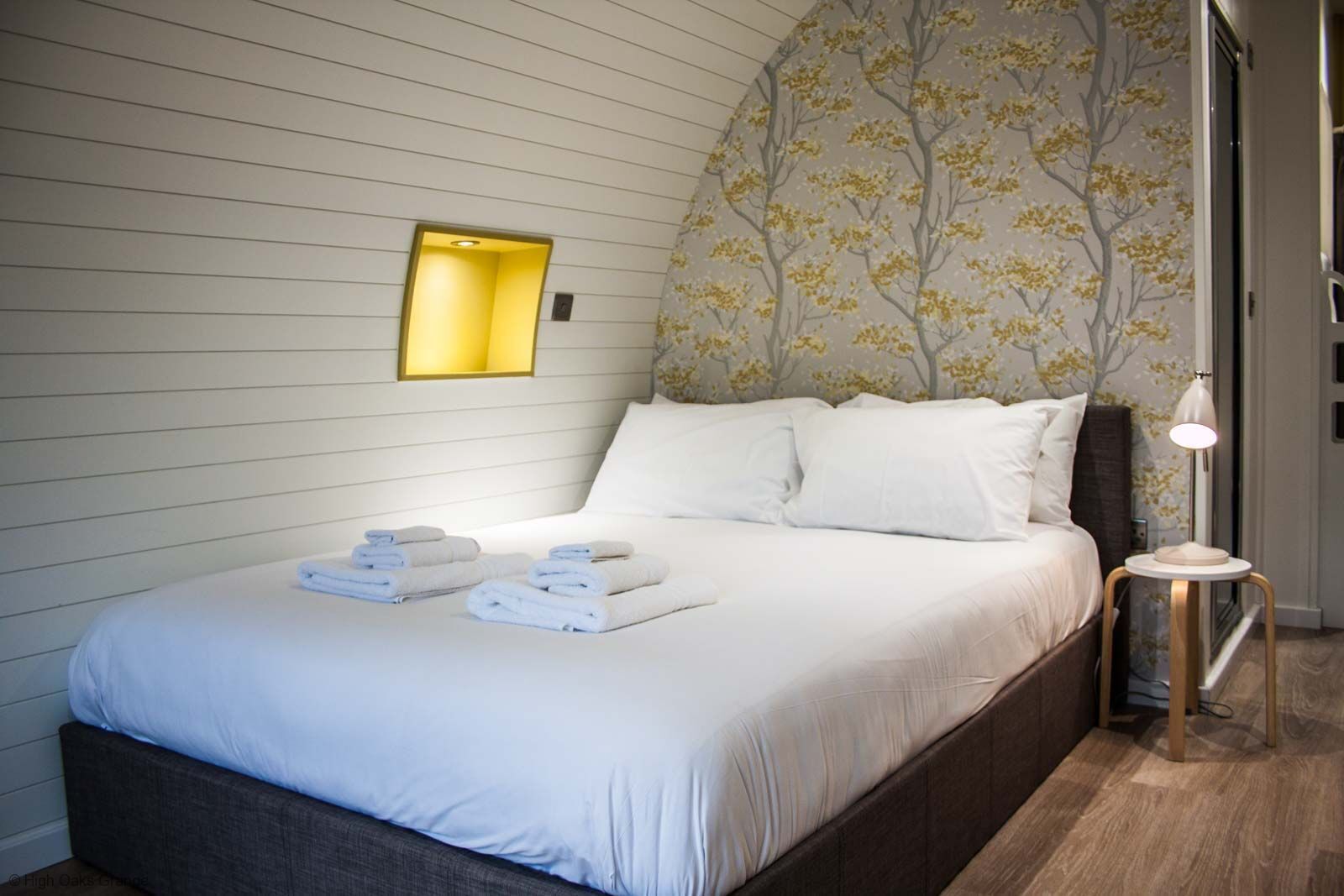 A neatly made bed with folded towels, a bedside lamp, and a wall with a tree pattern.