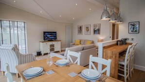 Un comedor con una mesa de madera dispuesta para cuatro personas, sillas blancas y un televisor al fondo.