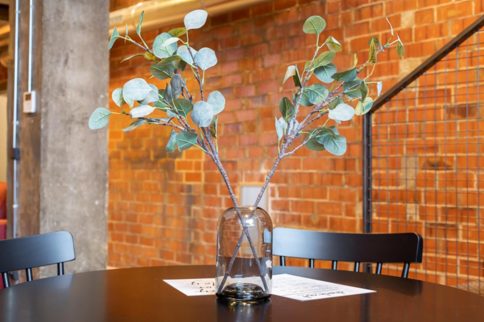 Une table sur laquelle est posé un vase en verre transparent contenant une plante aux feuilles vertes et aux fleurs blanches, le tout sur fond de mur en briques.