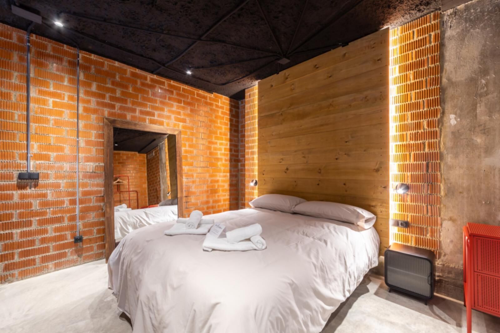 Une chambre d’hôtel moderne avec un mur en briques, un lambris en bois, un grand lit avec du linge de lit blanc et un radiateur rouge.