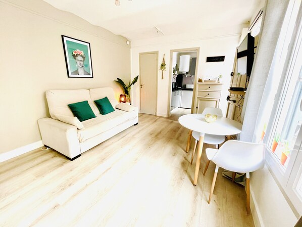 A living room with a white sofa, green cushions, a round table, and chairs. There is a kitchen visible through an open doorway.