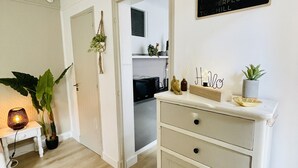 Un salón luminoso y minimalista, con suelo de madera, un mueble blanco con cajones, una mesita con lámpara y una planta en maceta.
