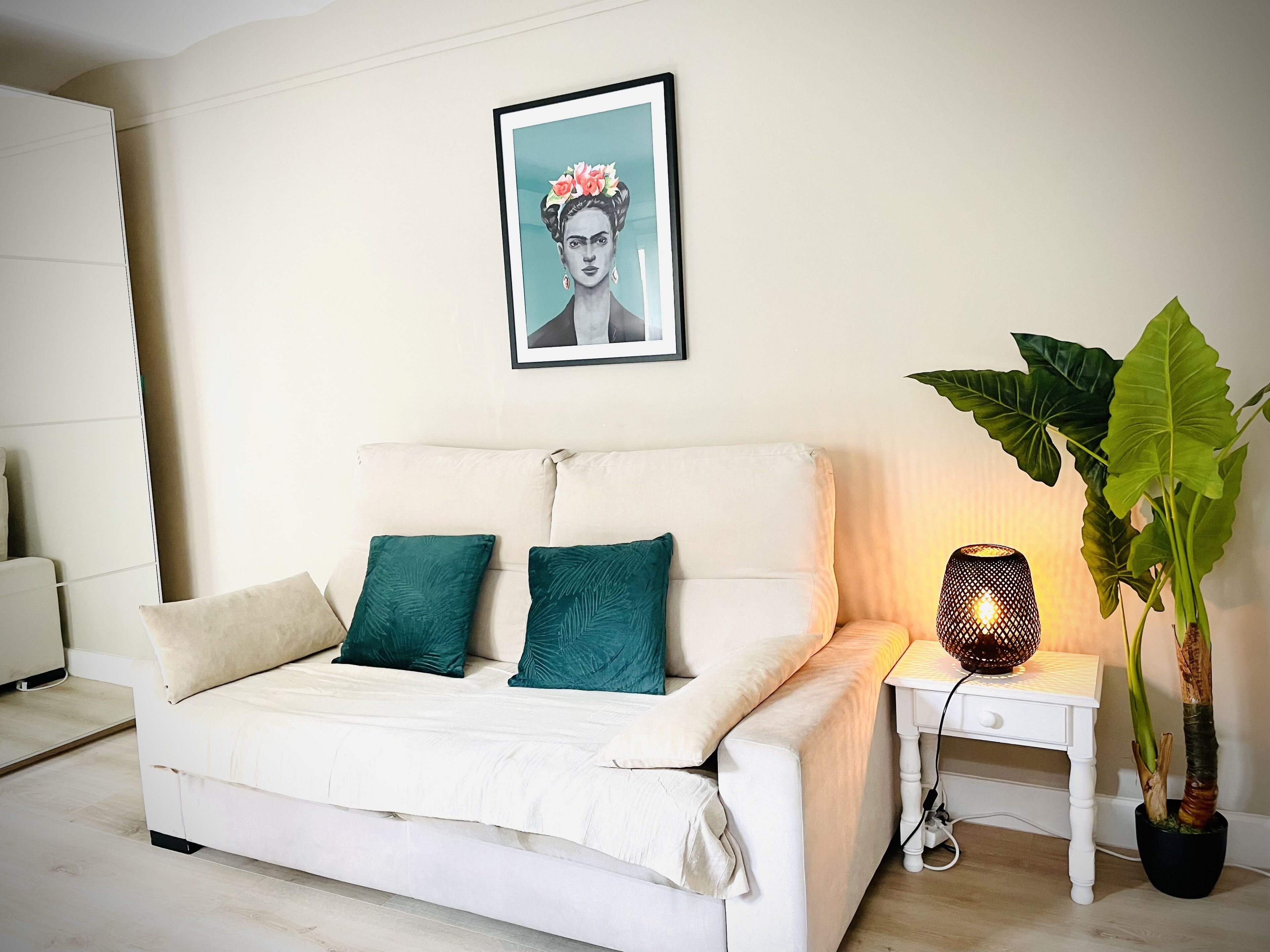 A bedroom with a white bed, teal pillows, a framed portrait on the wall, a bedside table with a lamp, and a potted plant.