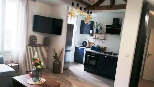 Een moderne woonkamer met een houten salontafel, een flatscreen televisie aan de muur en een keukenhoek met donkere kastjes en een fornuis.