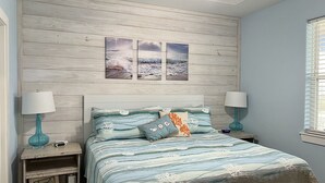 Une chambre à coucher avec une tête de lit en bois, deux tableaux accrochés au mur, un lit recouvert d’une courtepointe à rayures, deux lampes de chevet et une petite table de chevet avec une lampe.