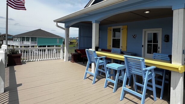 Une terrasse extérieure couverte avec une table jaune et des chaises bleues, une clôture blanche et un drapeau américain.