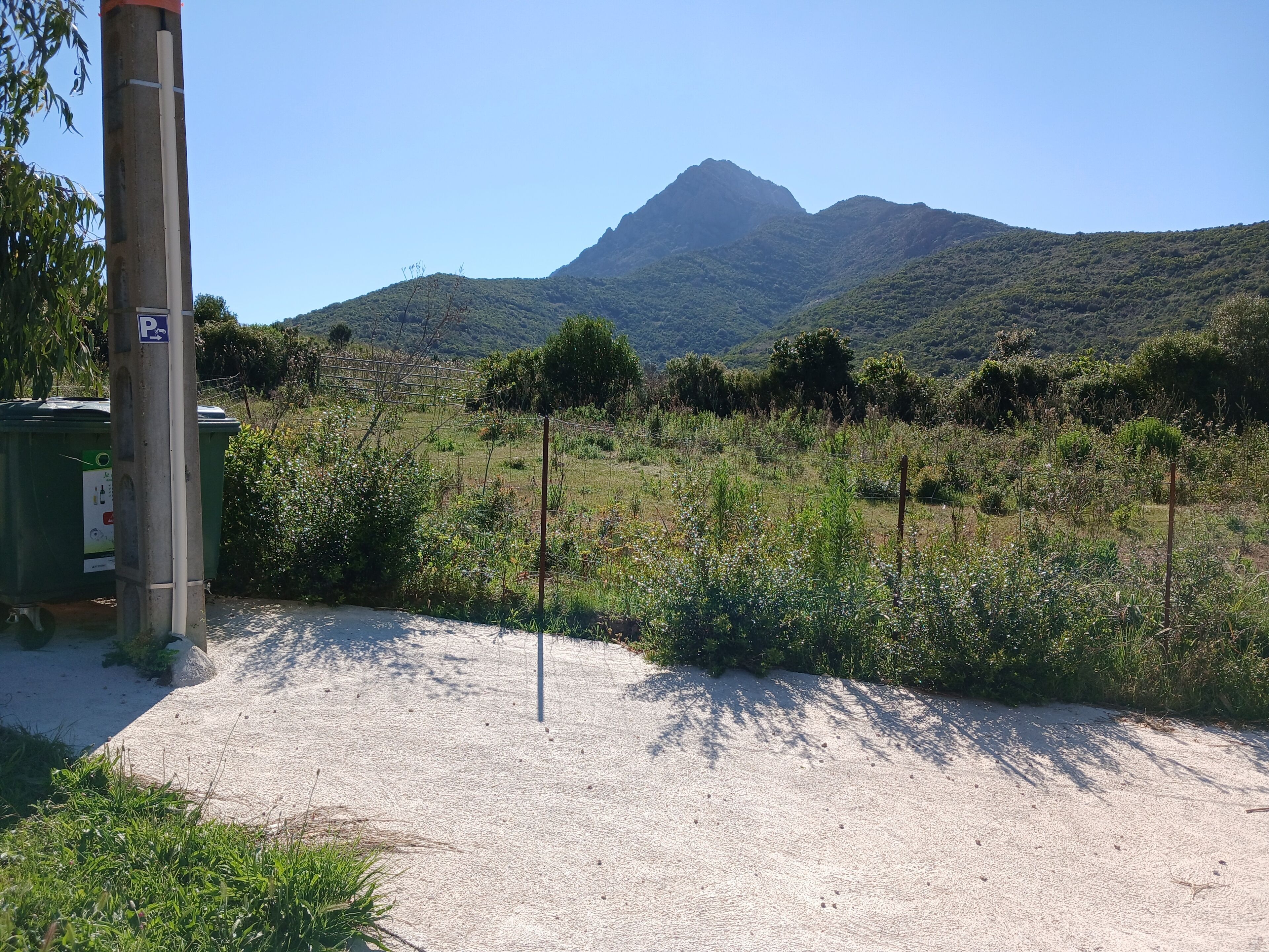 A parking area with a dirt path leading to a green structure, surrounded by vegetation and a mountain in the background.