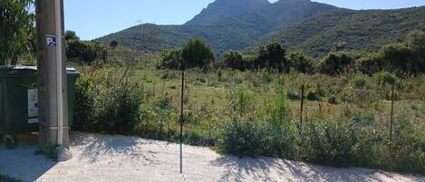 A parking area with a dirt path leading to a green structure, surrounded by vegetation and a mountain in the background.