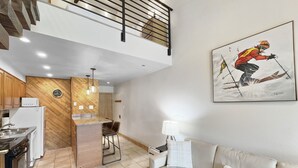 Un appartement moderne comprenant une cuisine, un coin repas et un salon. Il est doté d’un parquet en bois disposé en motif à chevrons, d’un canapé blanc et d’un tableau représentant un skieur.