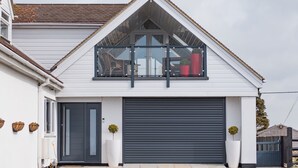 Une maison moderne avec une porte de garage sombre, un balcon doté d’une rambarde en verre et deux plantes en pot.