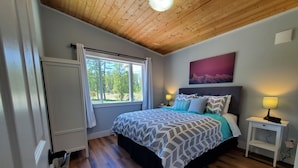 Quarto com teto de madeira, uma cama grande com roupa de cama estampada, uma mesa de cabeceira com abajur e uma janela com cortinas.