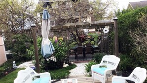 Eine Terrasse mit weißen Korbmöbeln, darunter zwei Stühle und ein kleiner Tisch, umgeben von Grünpflanzen und einem Zierzaun.