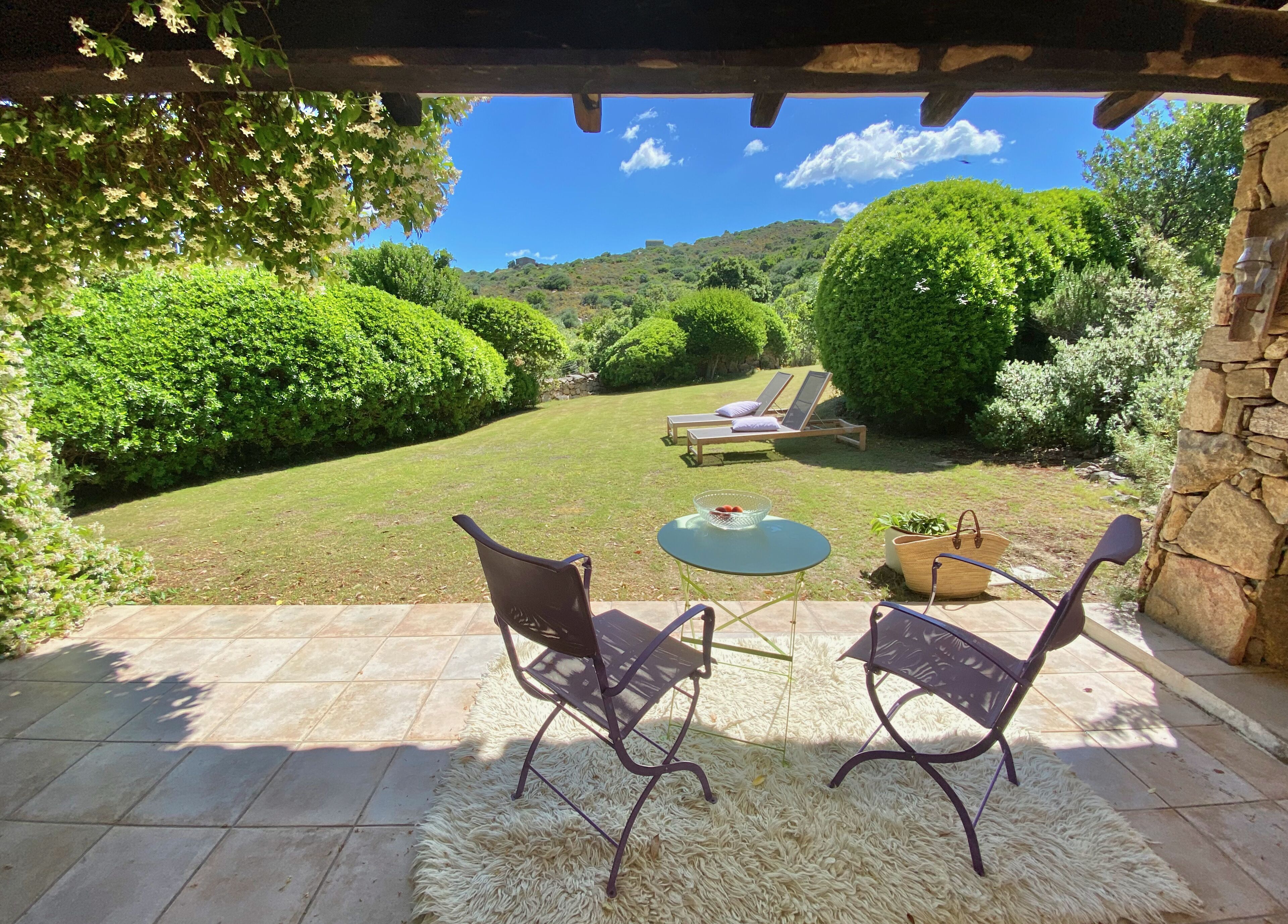 Eine Terrasse im Freien mit zwei Stühlen und einem Tisch, einer Steinmauer und Blick auf einen gepflegten Garten und Hügel im Hintergrund.