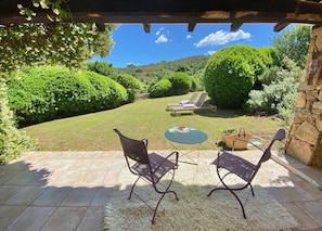 Un patio exterior con dos sillas y una mesa, un muro de piedra y vistas a un jardín cuidado y colinas al fondo.