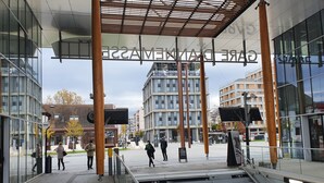 Una entrada moderna de edificio con fachadas de vidrio, un área peatonal cubierta y una escalera que conduce a un nivel superior.