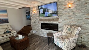 A living room with a flat-screen TV mounted on a stone wall, a fireplace below, a sofa, a patterned armchair, and a wooden coffee table.