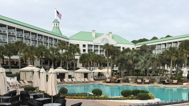 A hotel with a large pool surrounded by lounge chairs and umbrellas, a flag on top, and a green-roofed building. - Hilton Head with 4 rounds of golf and tennis a day included!!!! (Hilton Head Island)