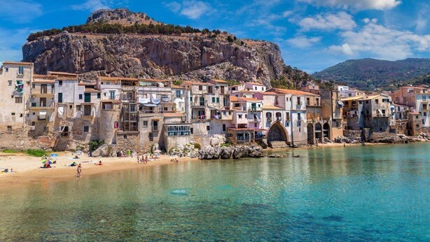 A coastal town with buildings along the waterfront, a prominent rocky hill in the background, and clear blue waters. - Villa Rosanna (Patti)