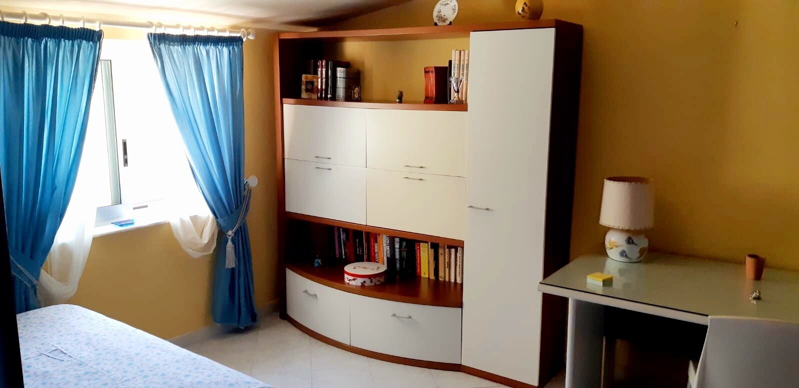Ein Zimmer mit einem Bett, einem Schreibtisch mit Lampe, einem Kleiderschrank und einem Fenster mit Vorhängen.