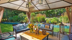 An outdoor patio with a covered seating area, a wooden table, and a view of a lush garden. - Hamptons Summer Rental - secluded pool + 5 minutes to beach (East Quogue)