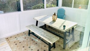 Une terrasse couverte avec une table et un banc, un ventilateur de plafond et une vue sur les arbres à travers les fenêtres.