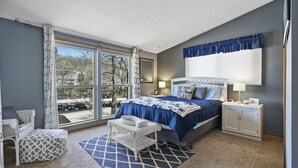 Un dormitorio con un ventanal grande, una cama con colcha azul, una mesita de noche blanca y una silla de mimbre.