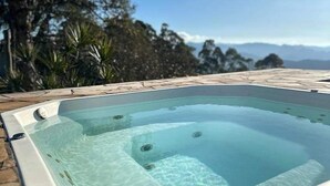 Uma banheira de hidromassagem retangular com revestimento de pedra, instalada ao ar livre sob um céu limpo e montanhas distantes ao fundo.